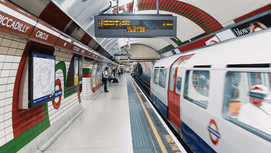 Interior view of a London Underground station platform with a white and red train arriving, tiled walls decorated with the London Underground roundel logo in red, white, and green, digital display boa