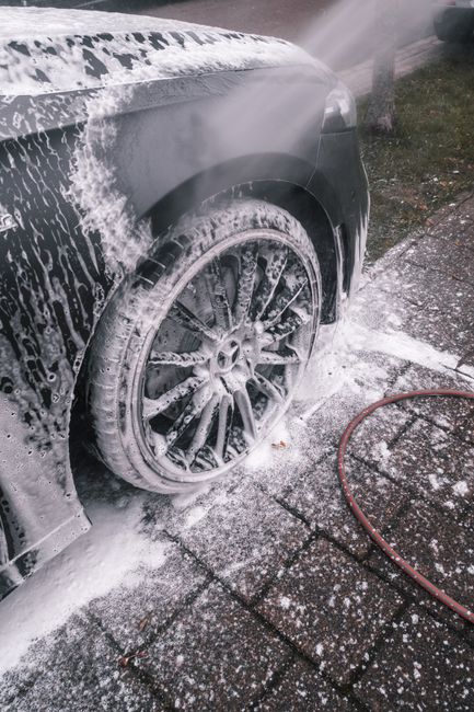 A close-up view of a black car being professionally cleaned at a commercial car wash facility. The vehicle's front wheel and tire are covered in white foam and soap suds from cleaning products, with s